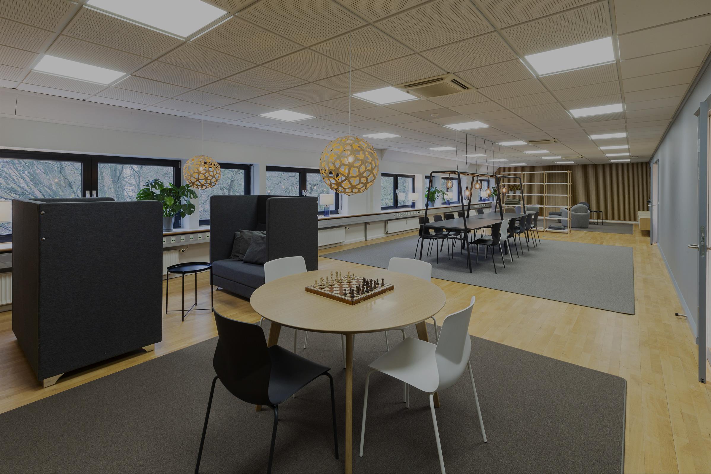 Lyst kontormiljø med loungeområde, mødebord, akustiske sofaer og skakbord i moderne skandinavisk kontorindretning.