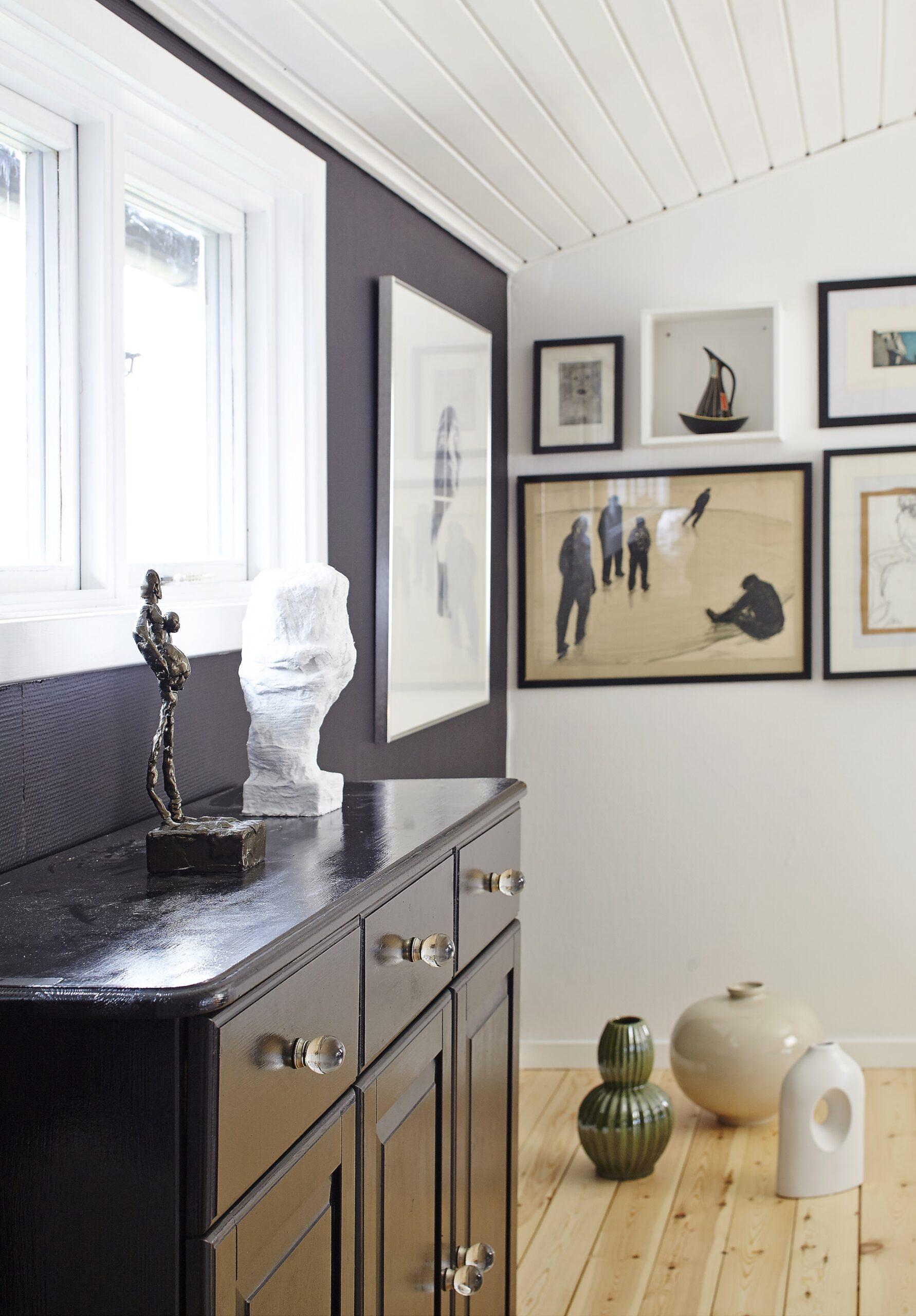 Sort skænk med små skulpturer foran væg med indrammet kunst og fotos i lyst og stilrent interiør.