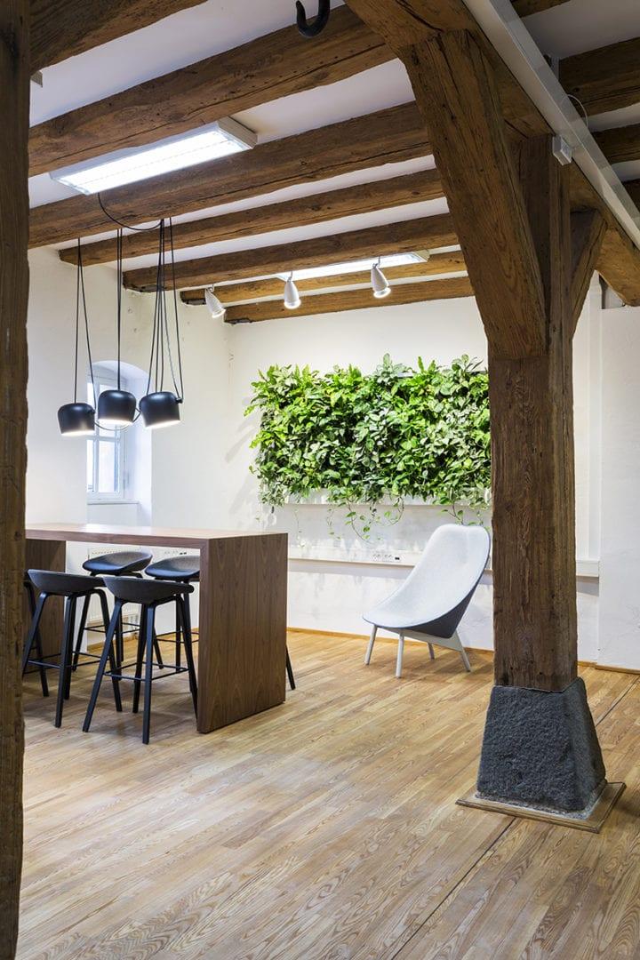 Moderne kontorindretning med synlige træbjælker, højbord, barstole og grøn plantevæg i skandinavisk stil.