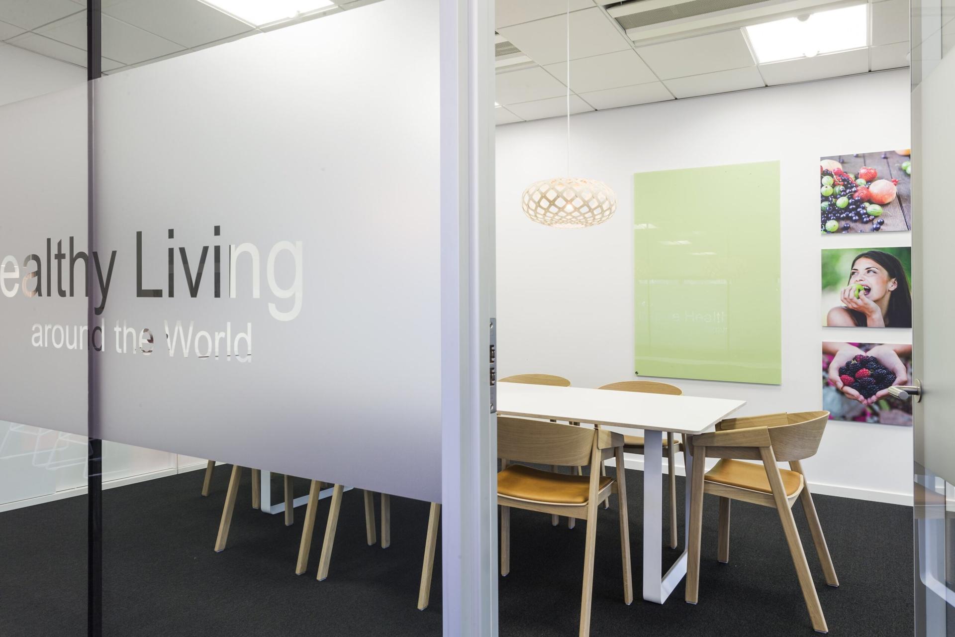 Mødelokale bag glasvæg med teksten “Healthy Living around the World”, bord, træstole og billeder af frugt.