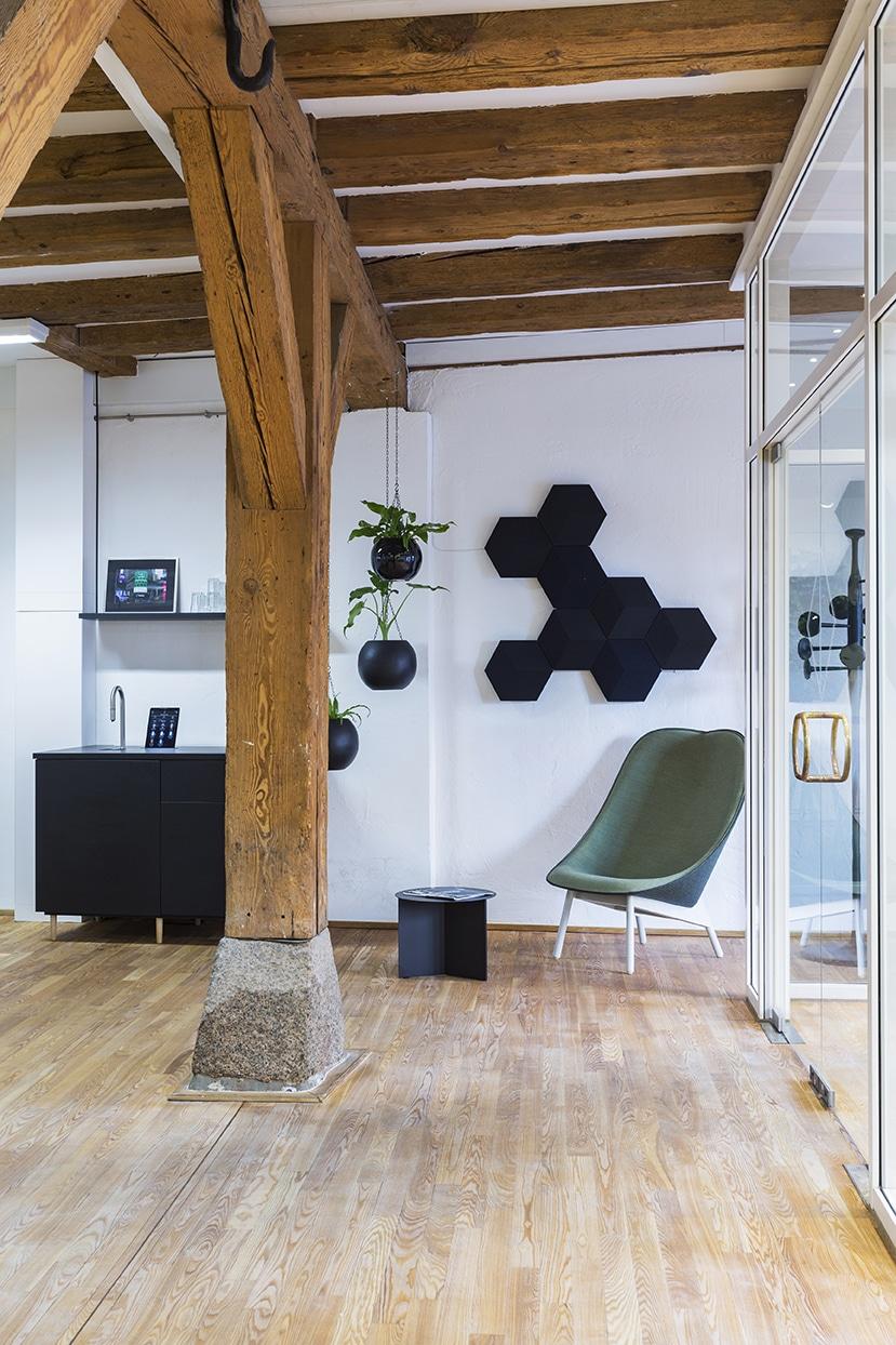 Moderne kontorindretning med synlige træbjælker, grøn loungestol, planter og geometriske akustikpaneler i skandinavisk stil.