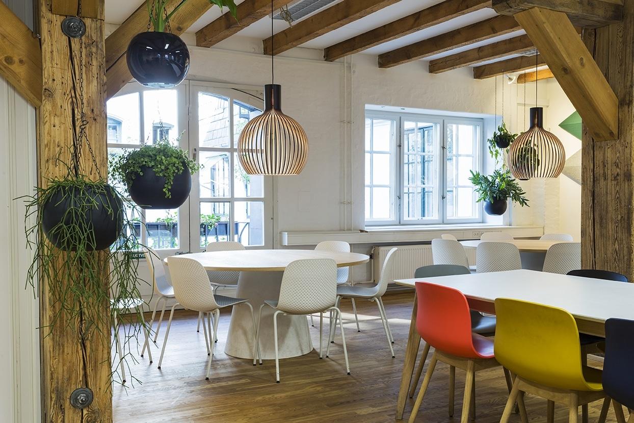 Lyst spiseområde med runde borde, hvide stole, træbjælker, hængende planter og dekorative pendellamper.