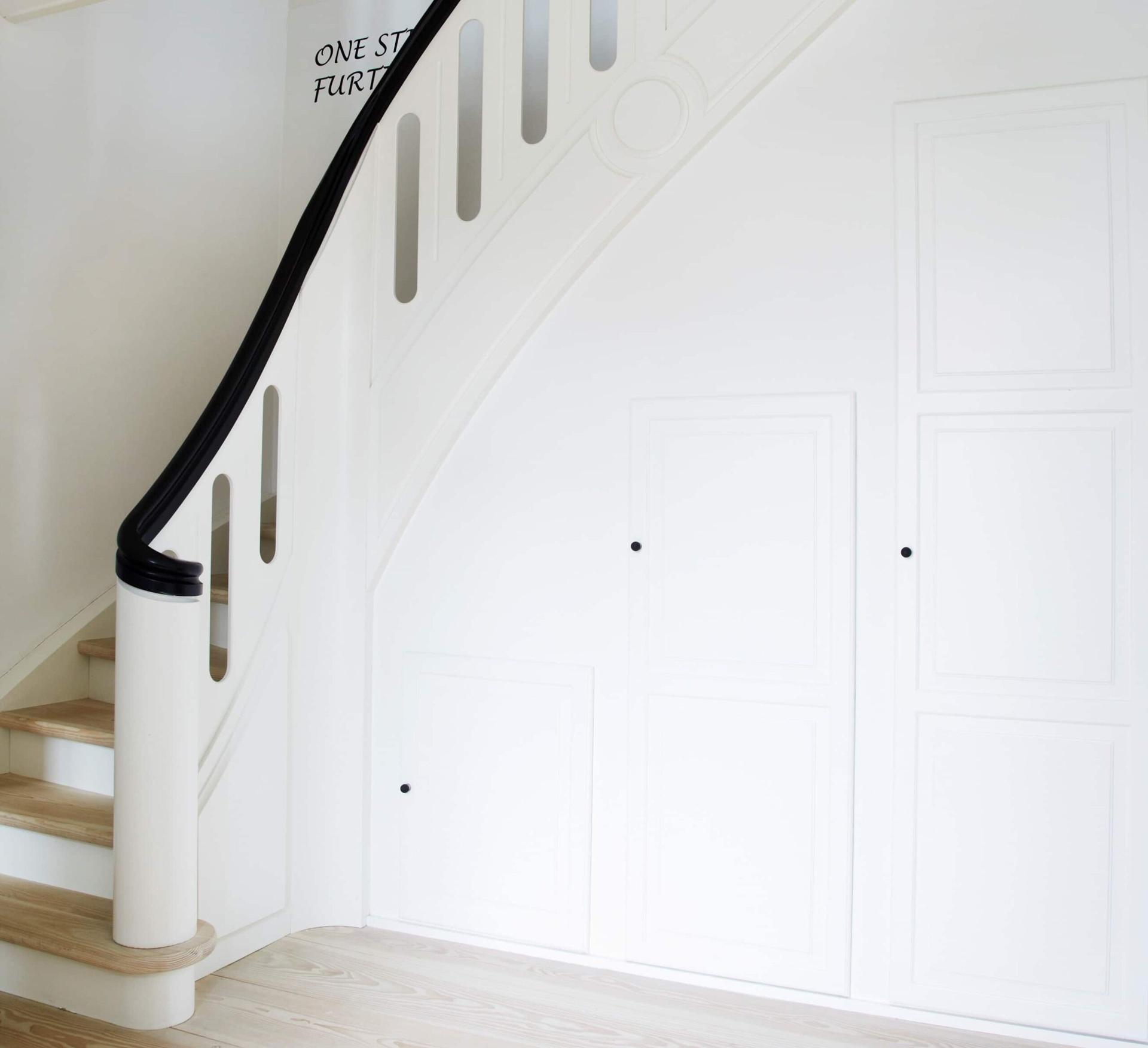 Hvid trappe med sort gelænder og indbyggede skabe under trappen i lyst og minimalistisk interiør.