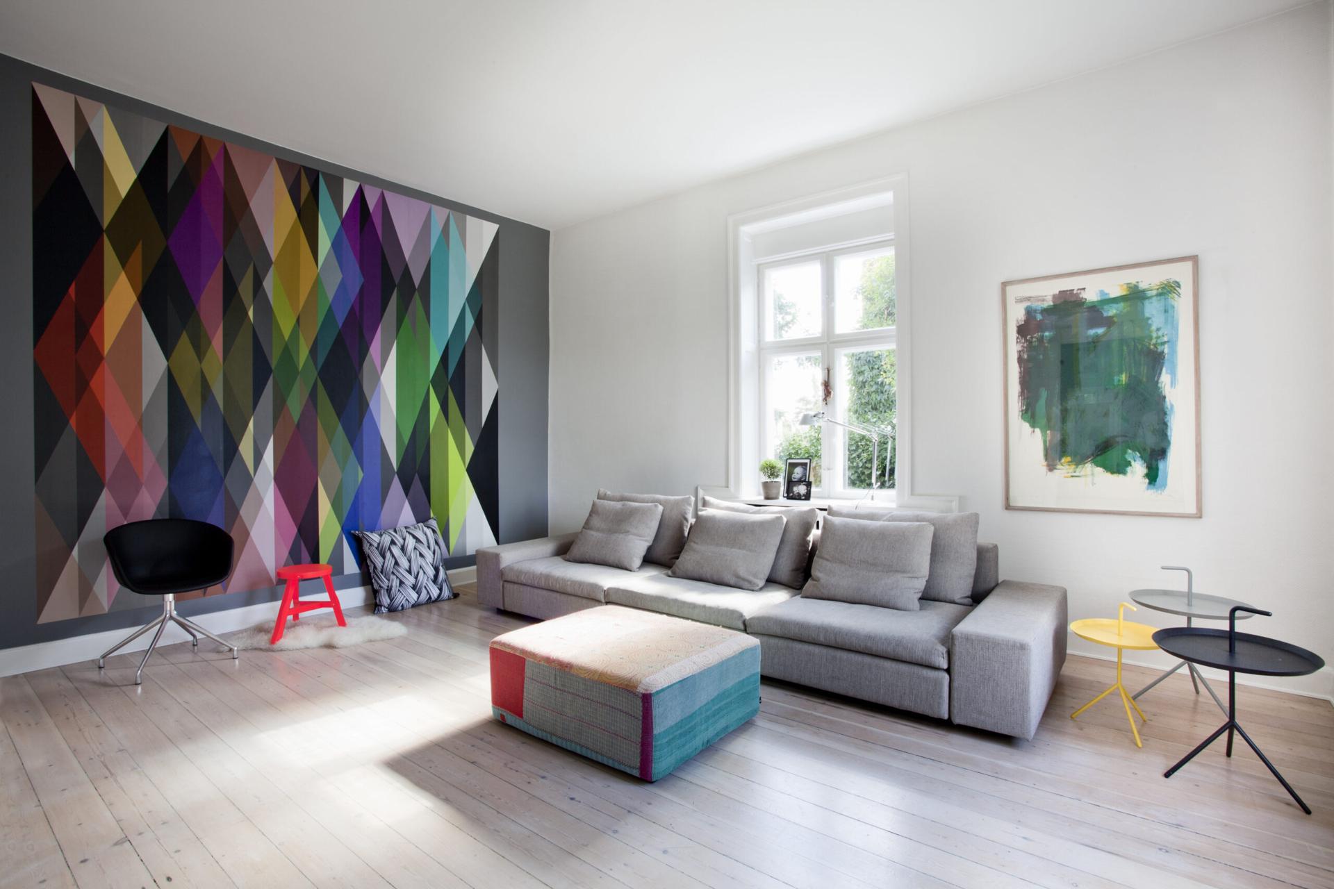 Lys stue med grå sofa, farverig geometrisk vægkunst, puf og moderne sideborde ved stort vindue.