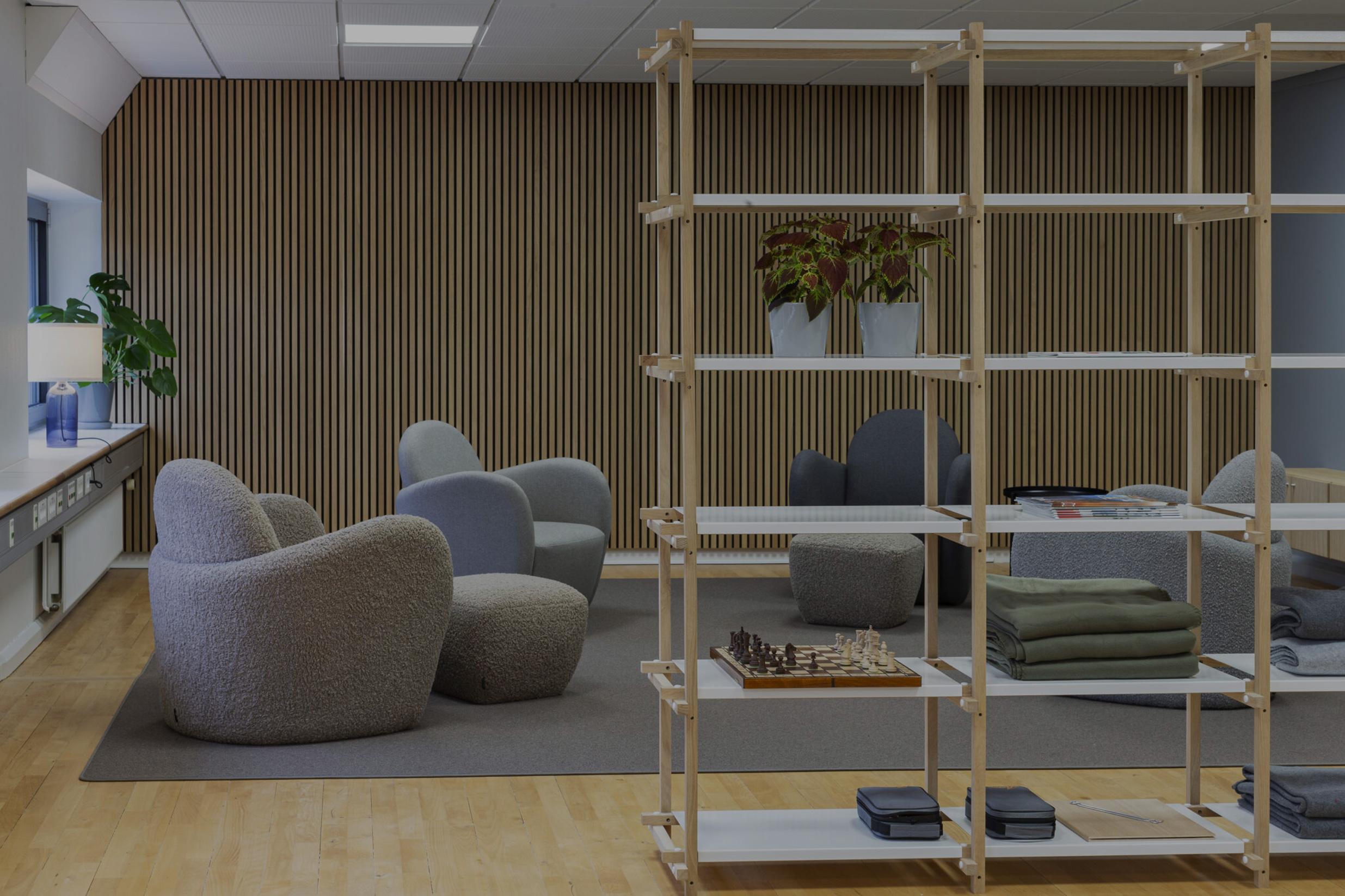 Moderne loungeområde med bløde lænestole, trælamelvæg, åbent reolsystem og planter i stilren skandinavisk kontorindretning.